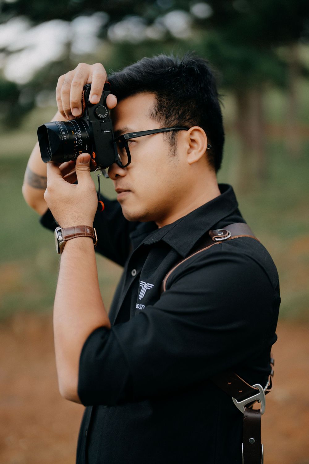 Young Asian man in black shirt photographing with DSLR camera outdoors, wearing glasses and leather camera strap.