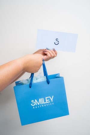 Hand holding a blue Smiley Aesthetics shopping bag with tissue paper and a branded card against white background.