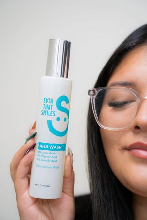 Woman with glasses holding Skin That Smiles AHA Wash bottle containing lactic, glycolic, and salicylic acid.