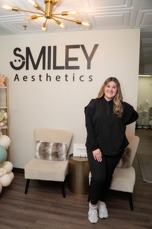 Woman in black outfit posing in front of Smiley Aesthetics sign in a modern, elegantly decorated medical spa.