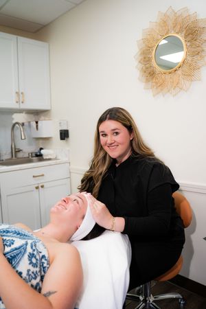 Esthetician smiling while performing a facial treatment on a client in a modern spa room.