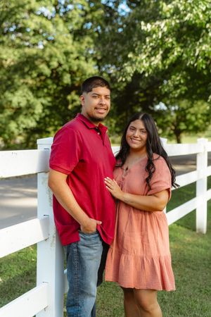 A couple in red and peach colored outfits pose together against a white fence in a sunny outdoor setting.