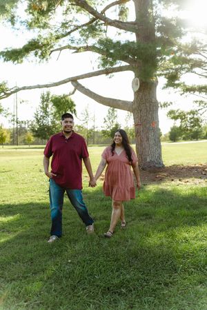 A couple stands together on a grassy hillside under tall trees during a sunny afternoon photo session.