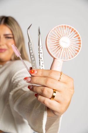 Close-up of hands holding beauty tools including tweezers and a pink mini fan.