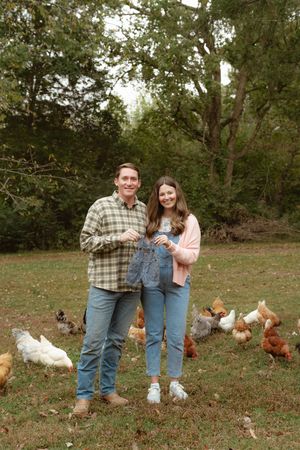 A couple stands in a grassy field surrounded by free-range chickens and hens on a sunny day.