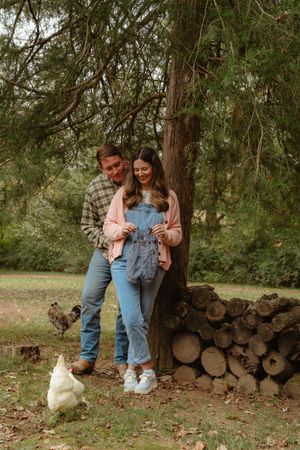 A couple stands by a woodpile in a rustic outdoor setting with chickens roaming nearby in the grass.