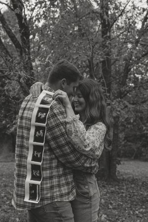 A couple shares an intimate moment outdoors while holding ultrasound photos in a wooded autumn setting.