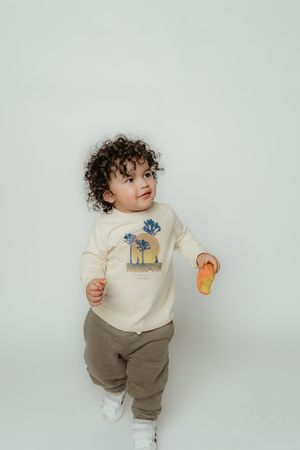Toddler in cream sweater with palm tree design playing with toy against white background.
