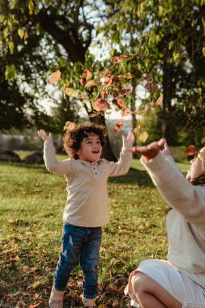 A toddler in jeans and white sweater plays joyfully in autumn leaves while someone watches nearby.