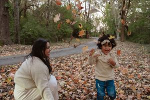 A person in casual attire playfully throws autumn leaves in the air while standing on a leaf-covered forest path.
