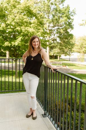 A woman in a black tank top and white pants poses casually on an outdoor railing in a sunny garden setting.