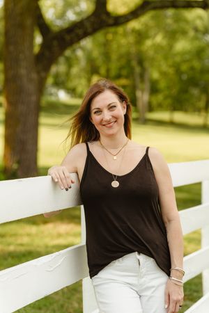 Woman in black tank top poses against white fence in outdoor setting with green trees in background.