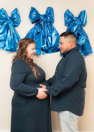 Pregnant couple holding hands smiling at each other with blue bow balloons on wall behind them.
