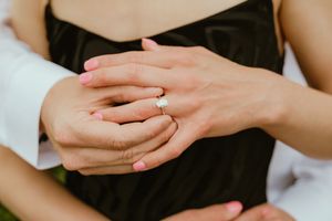 Close up of hands showing off an engagement ring with pink manicured nails against a black dress.