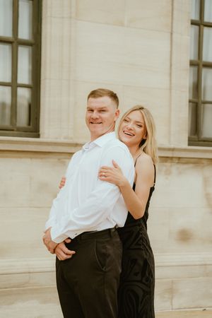 A couple in formal black and white attire share a joyful embrace and laugh together outside a stone building.