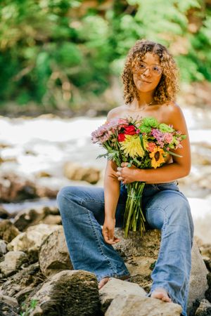 Person sitting on rocks by stream holding vibrant wildflower bouquet.