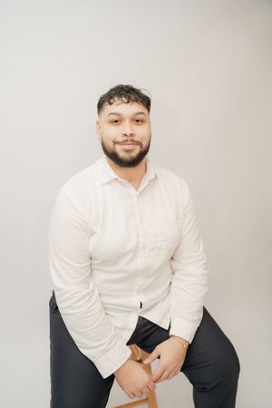 A young bearded man in a white button-up shirt sits relaxed on a wooden stool against a light gray background.