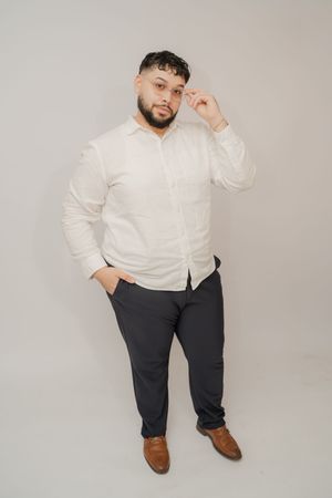 Man in white linen shirt and dark trousers posing confidently against a light gray background.