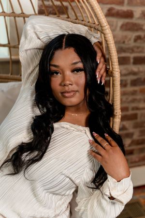 Portrait in cream sweater against brick wall with rattan chair detail.