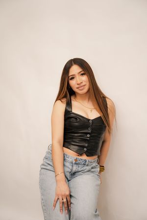 Young woman in black leather crop top and light wash jeans posing against a white background.