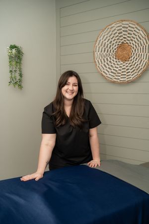 A smiling massage therapist in black scrubs stands beside a blue massage table in a serene treatment room.