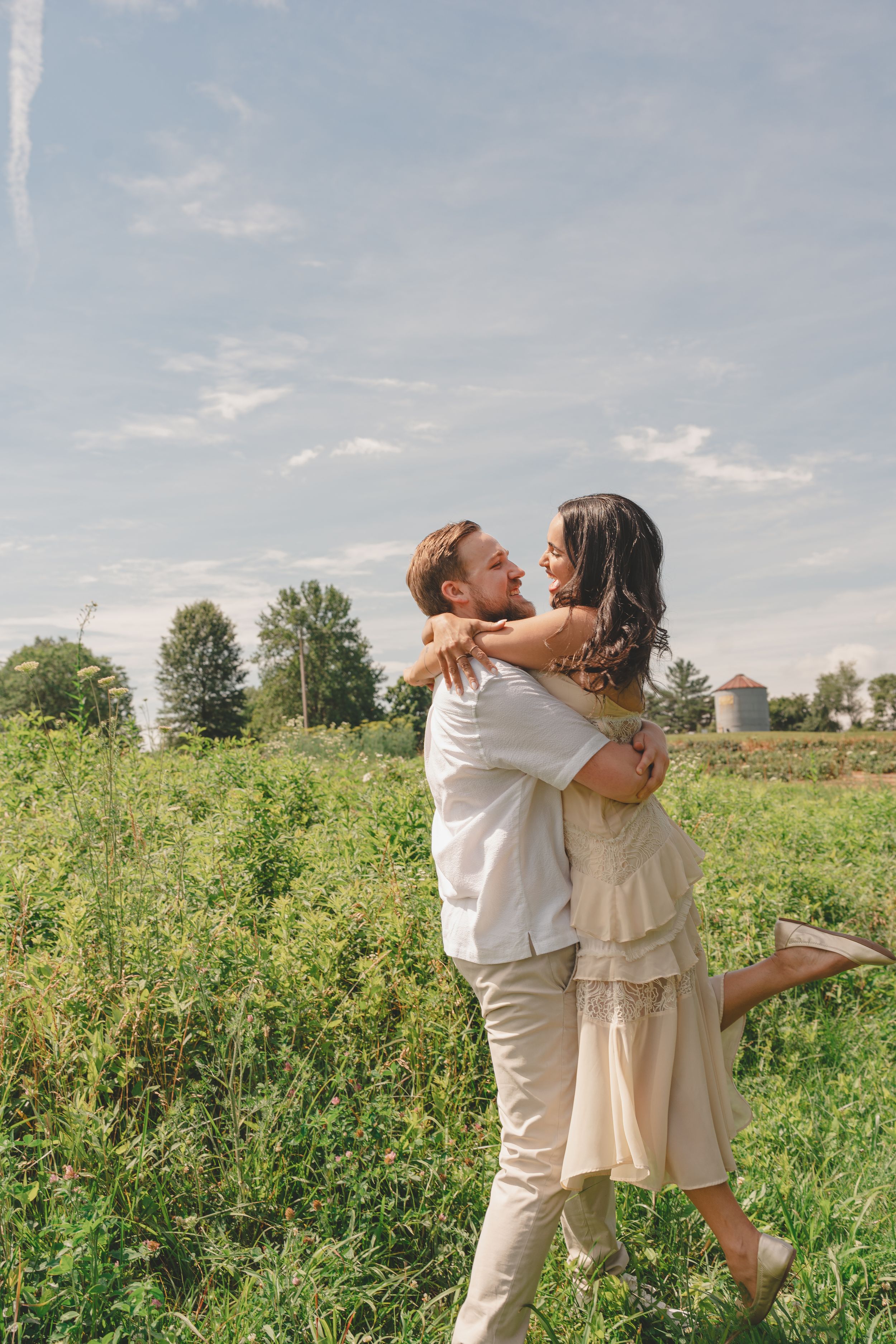 Couple embracing in a lush green field on a sunny day, woman in beige dress lifting her leg romantically.