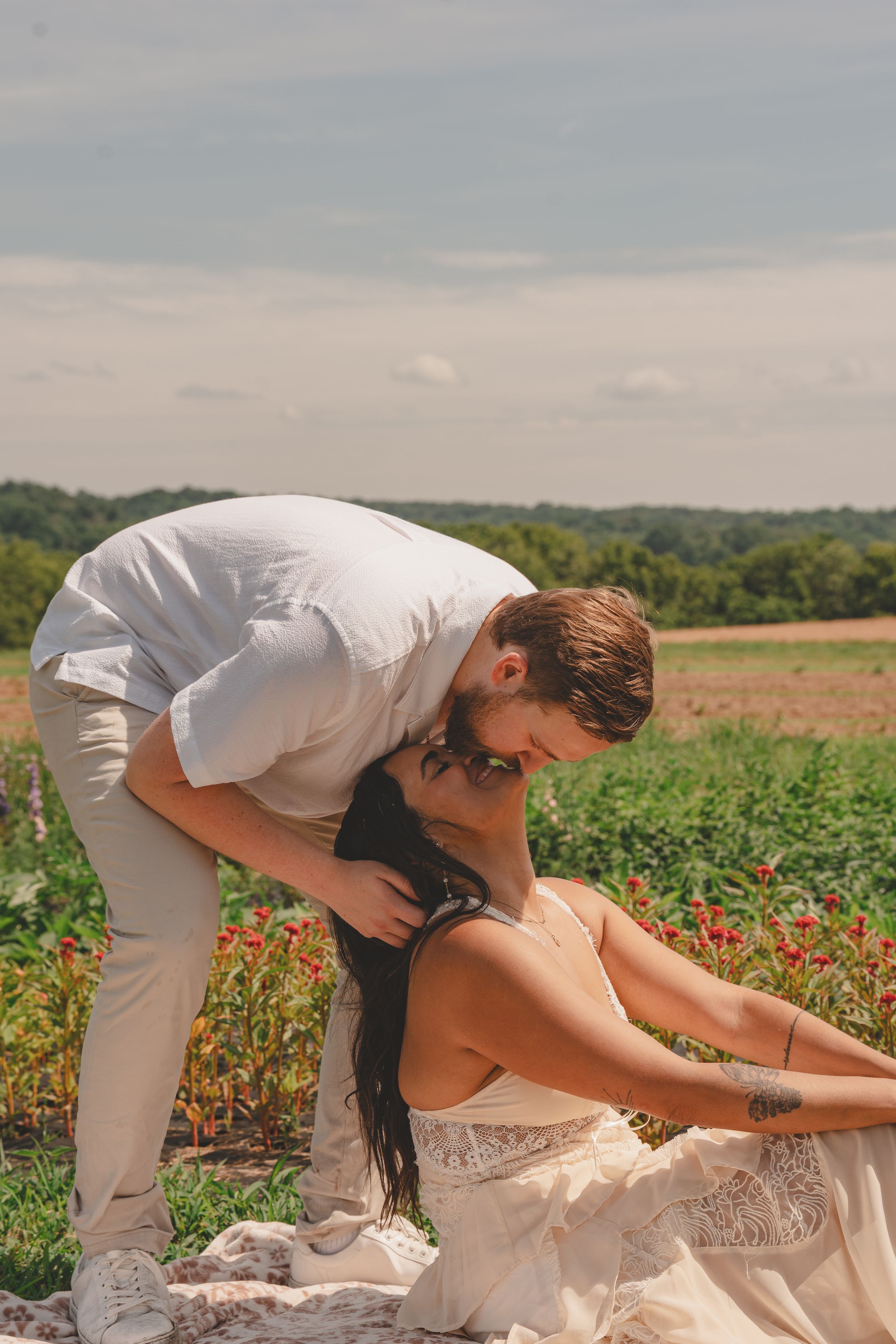 Romantic couple in white attire sharing a tender moment in a blooming field with green trees in the background.