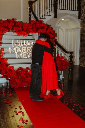 A marriage proposal setup with a red carpet walkway decorated with roses and illuminated 'Will You Marry Me' sign.