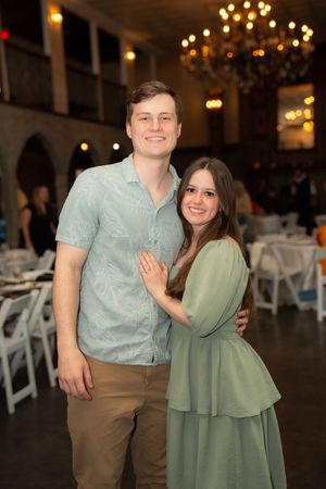 A smiling couple poses together at an elegant indoor event venue with chandeliers and white chairs in the background.