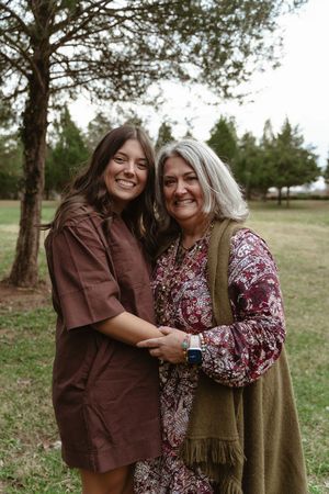 Two women smiling and embracing outdoors in a park, wearing brown and floral outfits on a cloudy day.