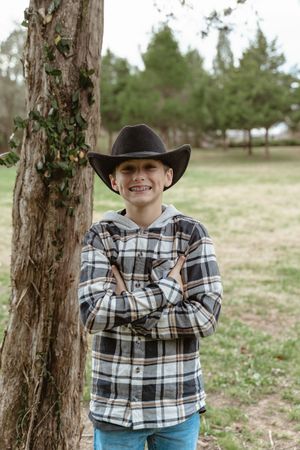 Smiling boy in a black cowboy hat and plaid flannel shirt stands with arms crossed beside a tree in a park.