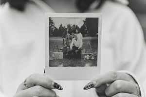 Hands holding a vintage Polaroid photo of a family group portrait in black and white outdoor setting.