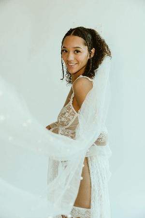 A person wearing a white lace robe and lingerie poses against a light background in an ethereal bridal boudoir photo.