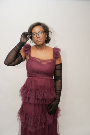 Woman in burgundy ruffled gown and black mesh gloves adjusting glasses, posing elegantly against white background.