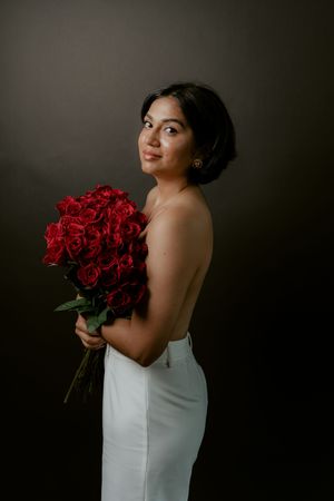 A person in a white dress holds a bouquet of vibrant red roses against a dark background in an artistic portrait series.