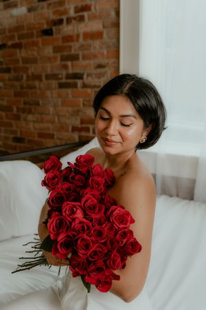 Woman in white dress holding vibrant red roses against exposed brick wall in romantic boudoir setting.