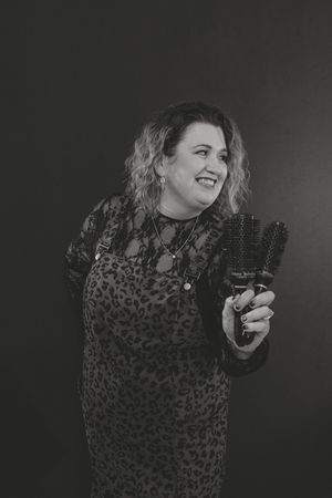 Smiling woman in leopard print dress holding hair brushes in a black and white studio portrait.