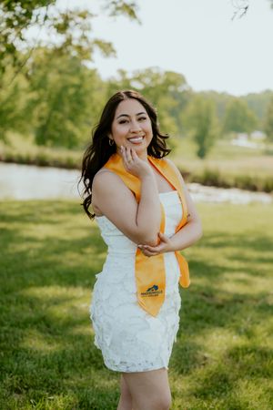 A person in a white lace dress and yellow sash poses playfully in a sunny outdoor setting.