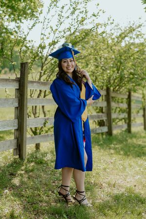 A graduate in blue cap and gown poses playfully in a sunny outdoor setting with white flowers and trees.