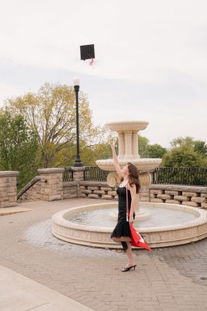 A graduate in a black dress joyfully tosses her cap near a stone fountain outdoors.