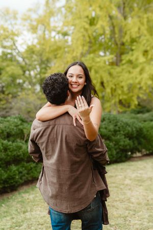 A couple shares a joyful embrace outdoors in a park with green foliage in the background.