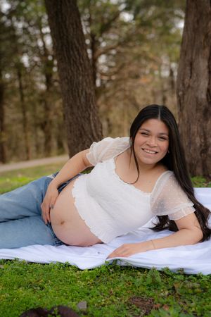Smiling pregnant woman lying on white blanket in park, wearing white crop top and jeans, cradling baby bump.