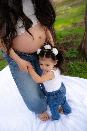 A toddler girl with white flower hair clips hugs her pregnant mother's belly outdoors on a white blanket.