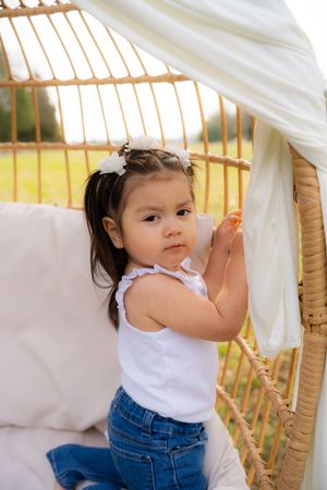 Toddler girl with pigtails in white top and jeans sitting in a wicker egg chair outdoors.