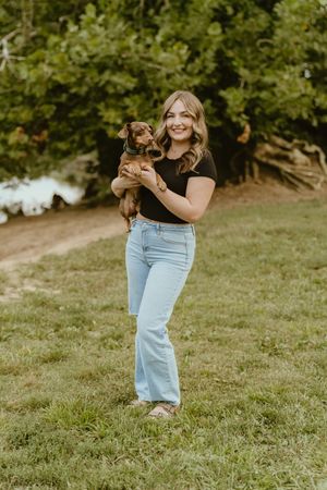 Person in light blue jeans holds and plays with small brown dog on grassy hillside in outdoor setting.