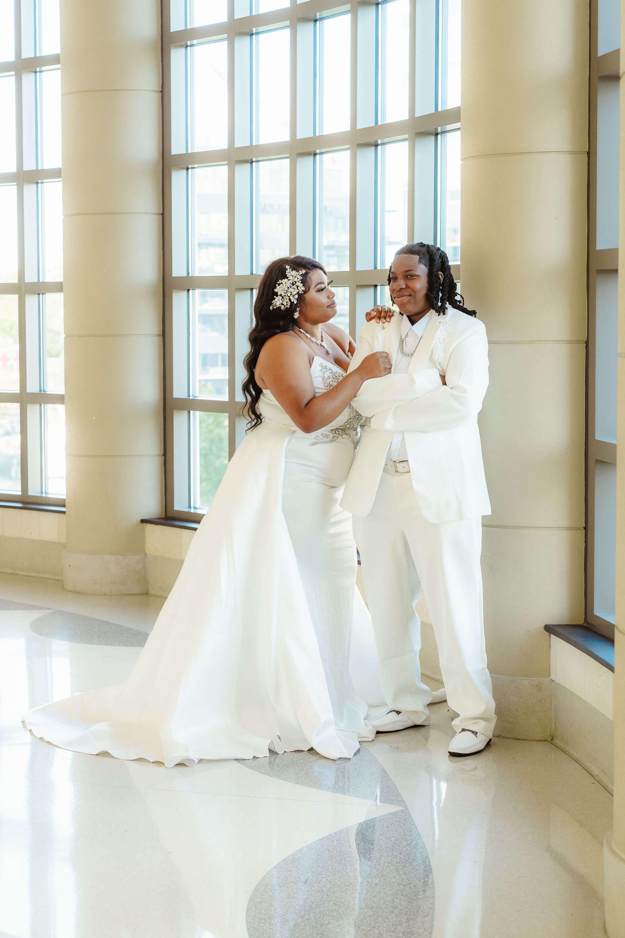 A romantic wedding couple in matching white attire share a tender moment by large windows in an elegant venue.