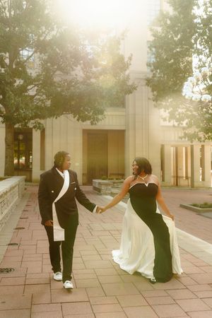 A couple in formal wedding attire dances playfully together on a stone patio with sunlit trees in the background.