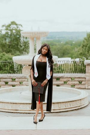 Elegant figure in a black dress and flowing cardigan poses gracefully on a stone terrace with scenic views.