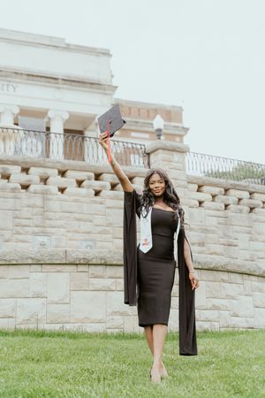 A graduate in a black dress and graduation gown poses for celebratory photos on a grassy lawn near stone walls.