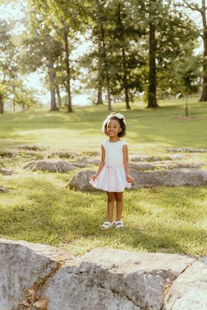 A young child in a white dress stands on a rock formation in a sunlit grassy park setting.